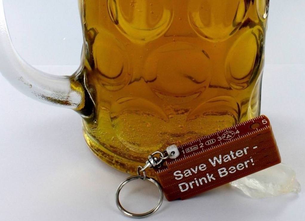 Kleiner bedruckter Zollstock 'Save Water - Drink Beer' mit Bierkrug im Hintergrund.