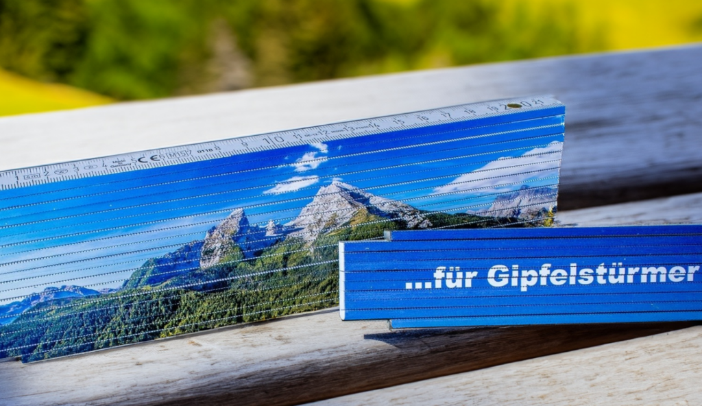Zwei Zollstöcke mit Werbedruck liegen auf einer hölzernen Unterlage. Ein Zollstock zeigt ein Bild einer Berglandschaft und der andere auf blauem Hintergrund die Aufschrift '...für Gipfelstürmer'.